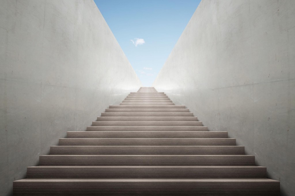 A wide staircase rising between light gray walls toward a bright sky, evoking steady forward progress