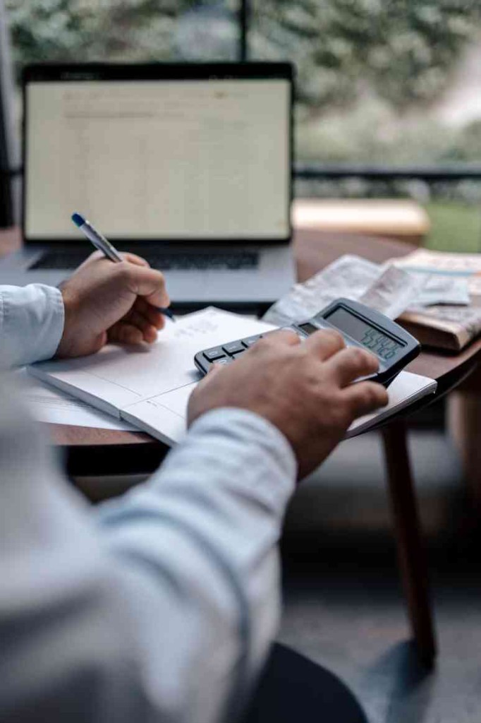 Person using a calculator and writing in a notebook while working on invoices at a desk