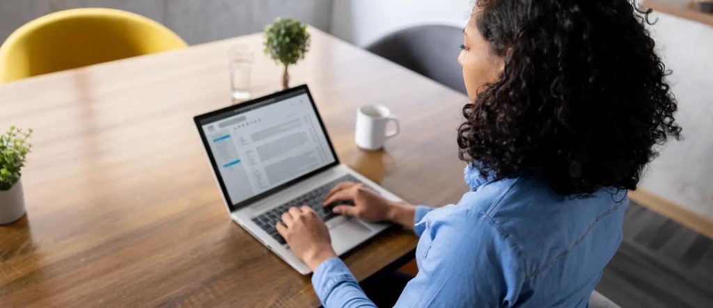Woman working at a wooden desk on a laptop with an email inbox on screen, coffee mug and plants nearby