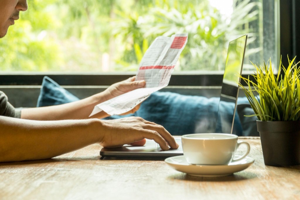Person at a wooden desk reviewing a paper invoice while using a laptop, with coffee and a plant nearby
